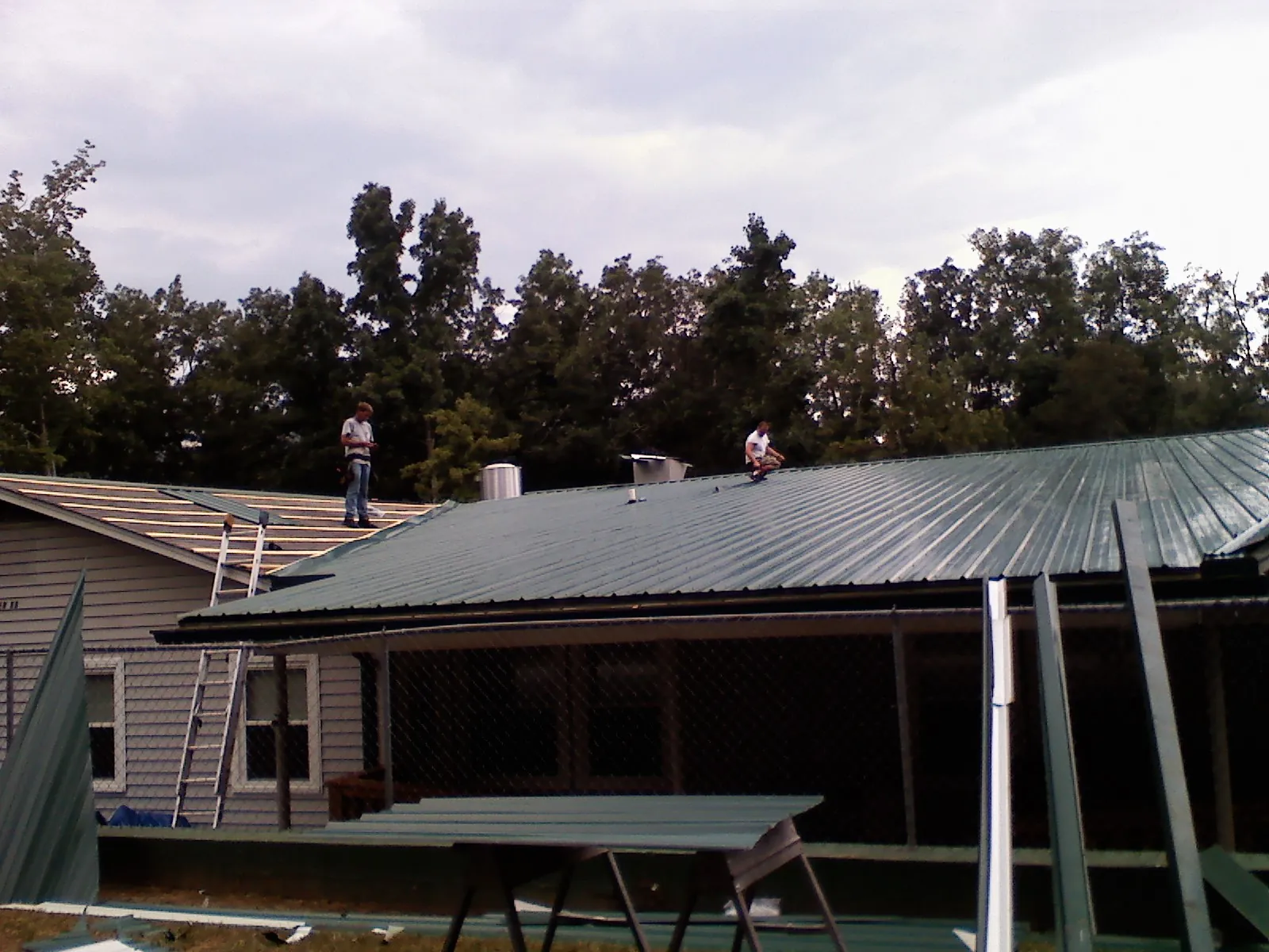 Two roofers installing green metal panels above a covered porch in Monroe